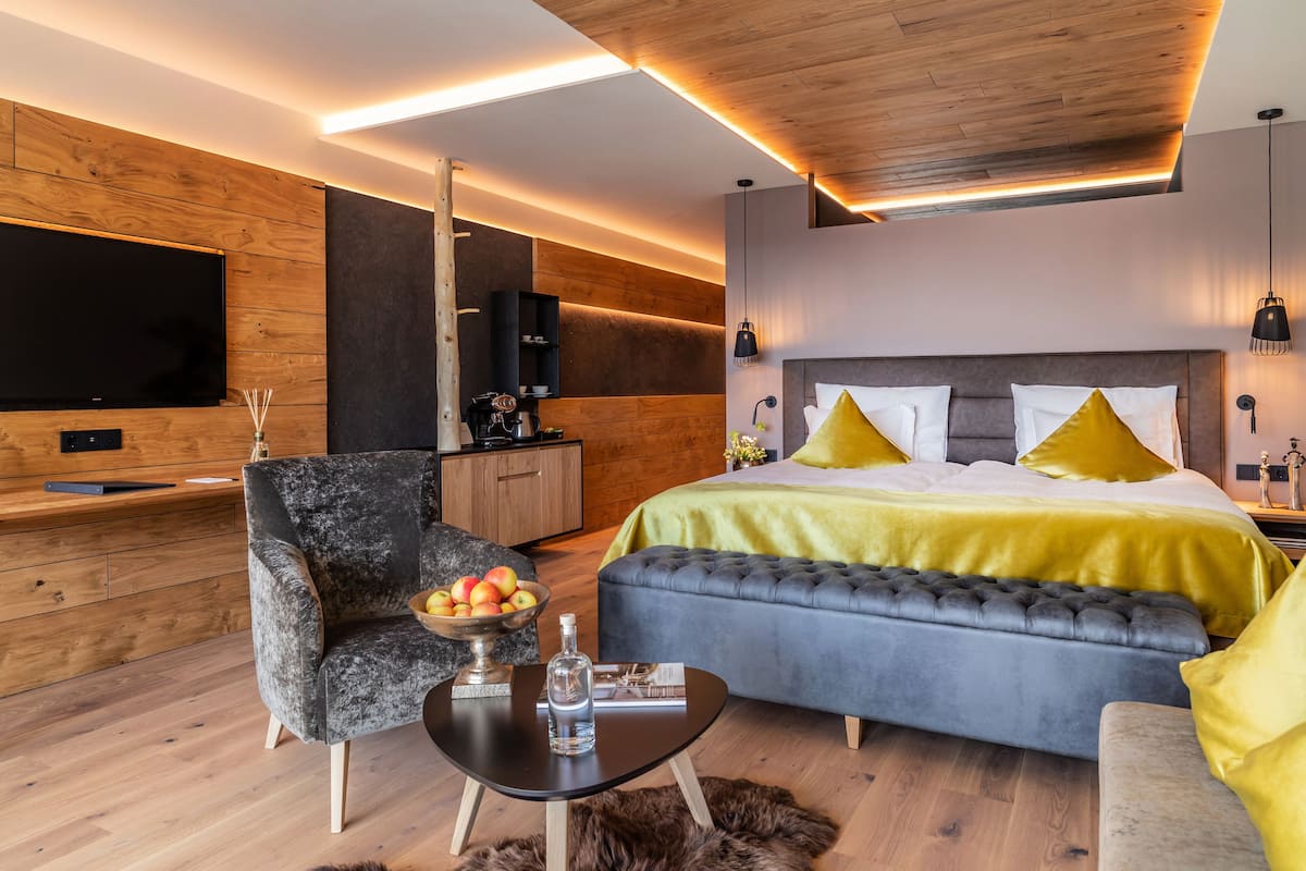 Una habitación de hotel moderna con techo de madera, una cama grande con almohadas amarillas, un sofá gris, un escritorio de madera con un televisor y una pequeña mesa con un cuenco de fruta.