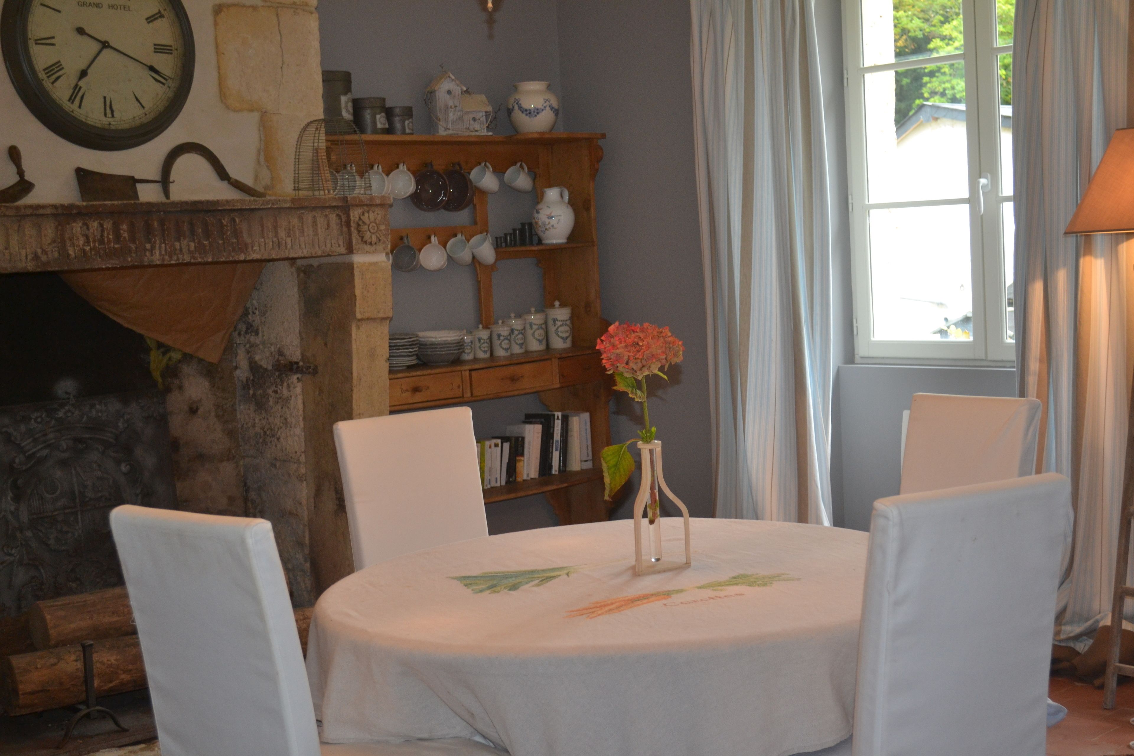 A dining area with a round table, white chairs, a vase with flowers, a clock, and a shelf with dishes and books.