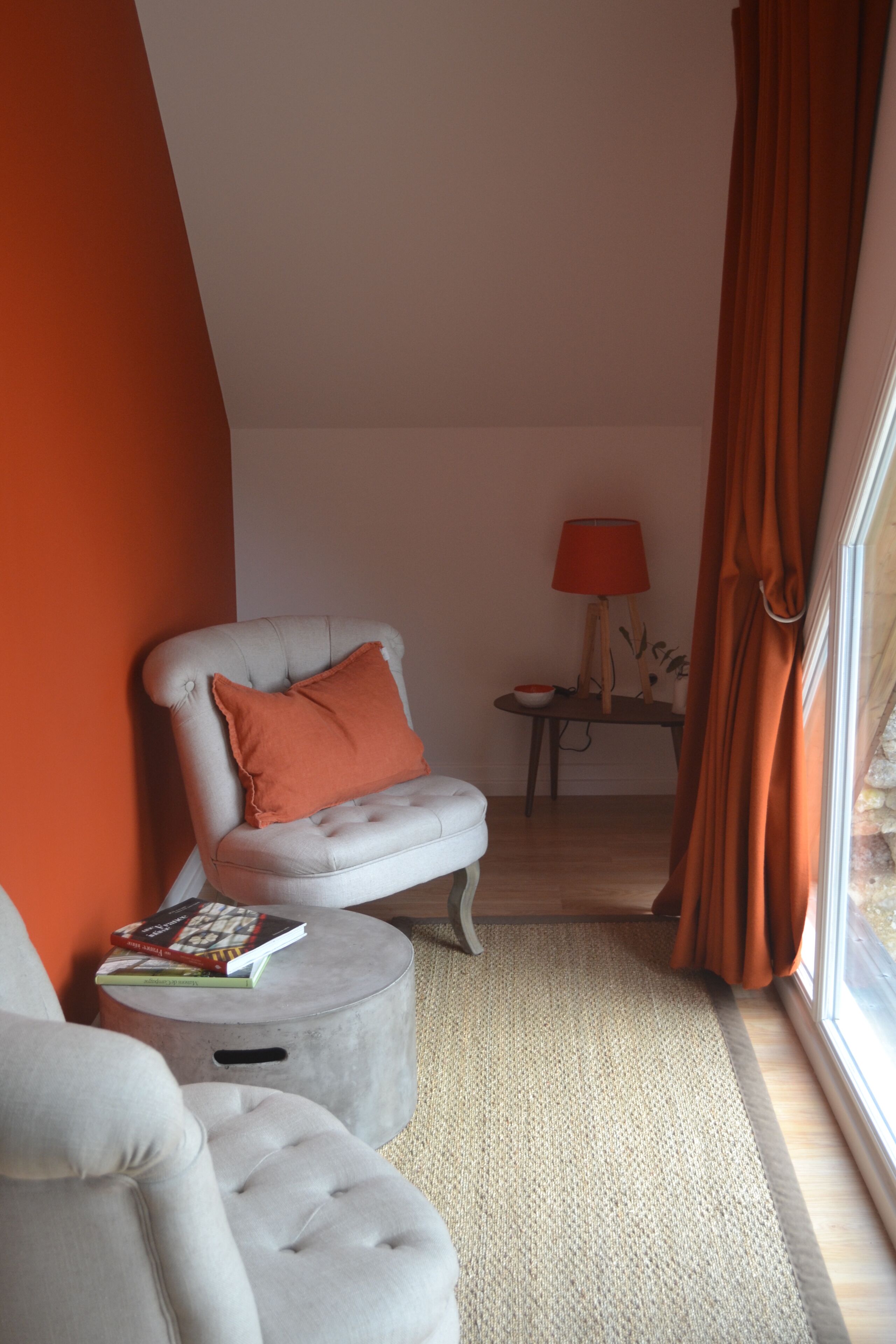 Une pièce avec un fauteuil blanc, un pouf rond, une petite table avec une lampe, et une vue par une fenêtre ornée de rideaux orange.