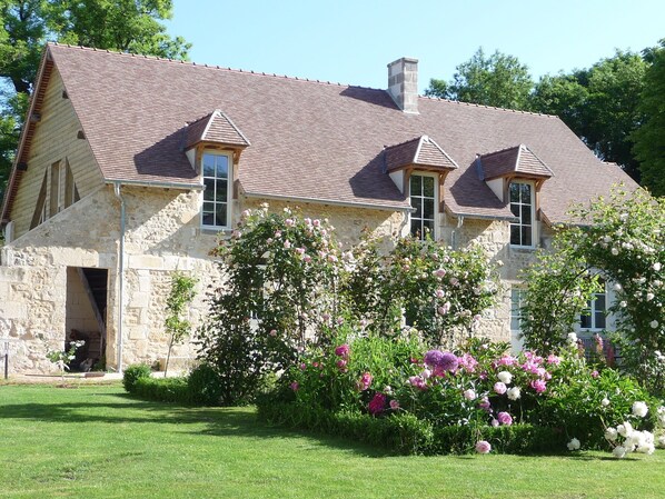 A stone house with a tiled roof, a large garden with flowering plants, and a clear sky.