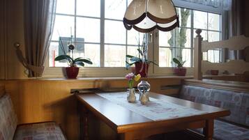 Un comedor acogedor con mesa de madera y banco, plantas en macetas y un amplio ventanal con cortinas.