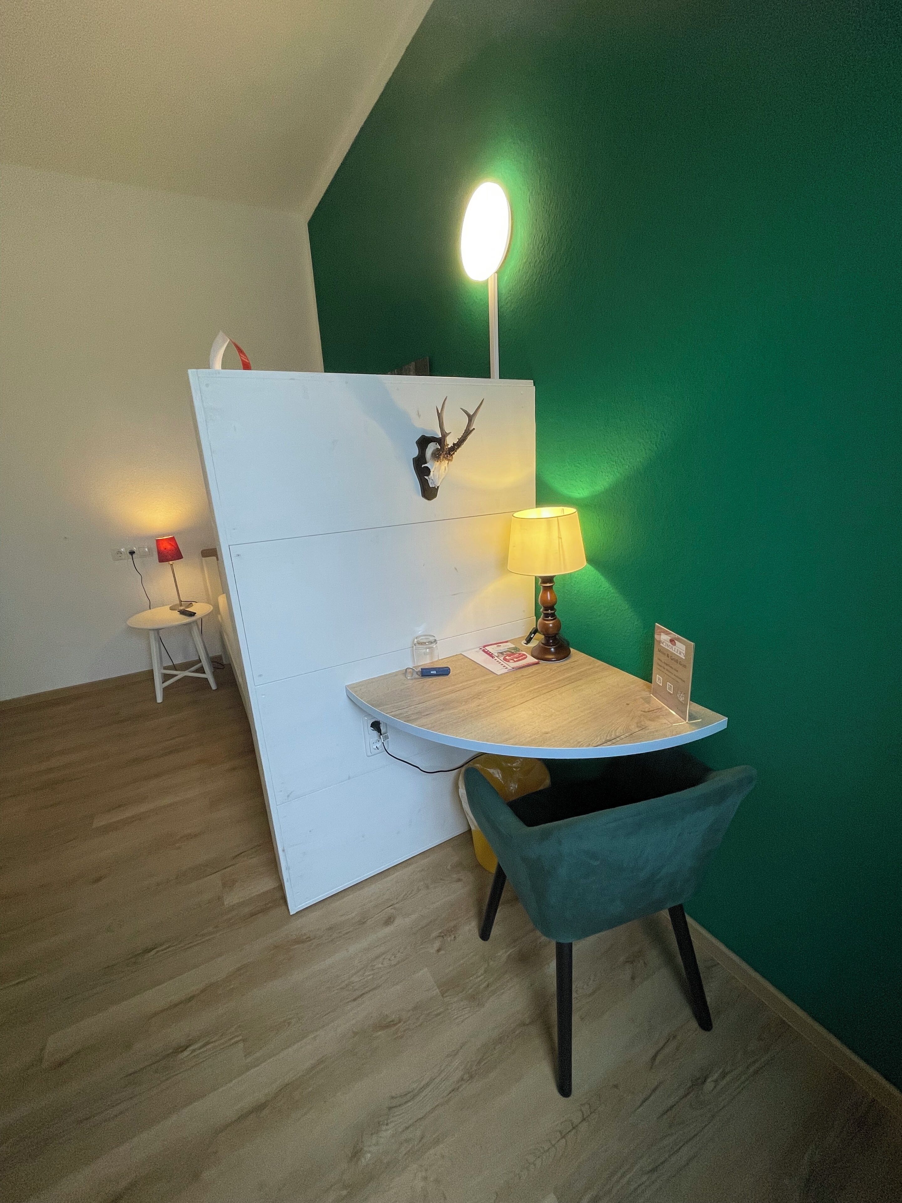 Ein Zimmer mit grüner Wand, einer weißen Trennwand, einem runden Tisch mit Lampe, einem Stuhl und einem kleinen Tisch mit roter Lampe.