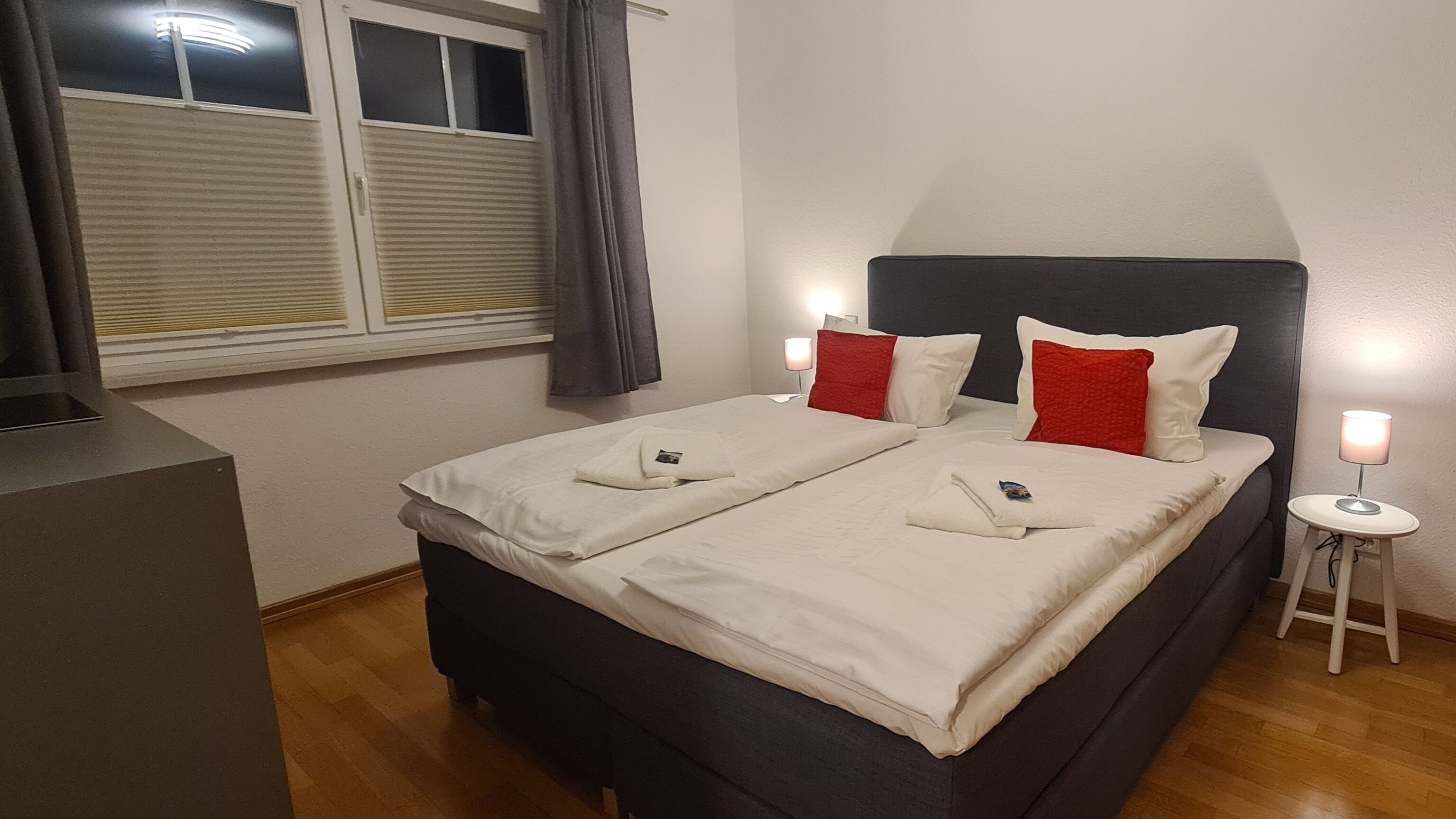 Ein Doppelbett mit weißen und roten Kissen, ein Nachttisch mit Lampe sowie ein Fenster mit Jalousien.