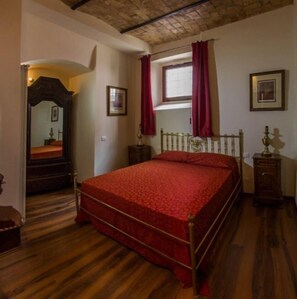 A bedroom with a wooden floor, a bed with a red bedspread, a nightstand, a dresser, a window with curtains, and a mirror. - Legacy (Sulmona)
