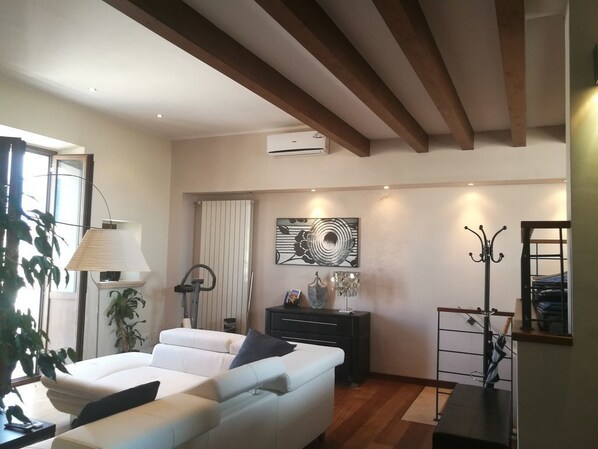 A modern living room with a white sofa, a black side table, and a floor lamp. There is a painting on the wall and a window with curtains. - Legacy (Sulmona)