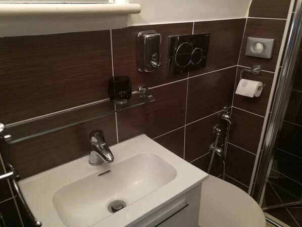 Un baño con un lavamanos blanco, un estante de vidrio, un dispensador de jabón negro, un porta papel higiénico y una zona de ducha.