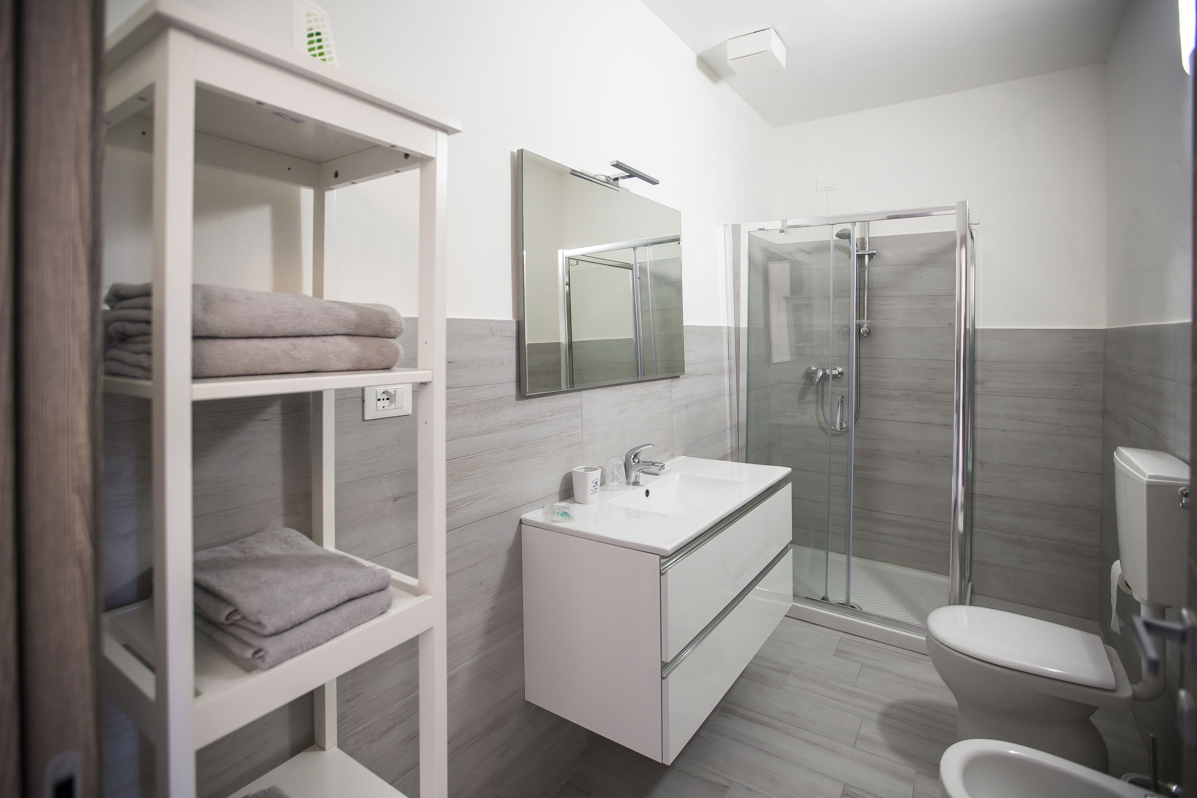 Un baño moderno con cabina de ducha de vidrio, un lavabo blanco y un estante empotrado con toallas dobladas.
