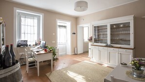 Une pièce bien éclairée, avec un bureau, un ordinateur et des bouteilles de vin. On y trouve également un meuble contenant de la verrerie et une plante en pot.