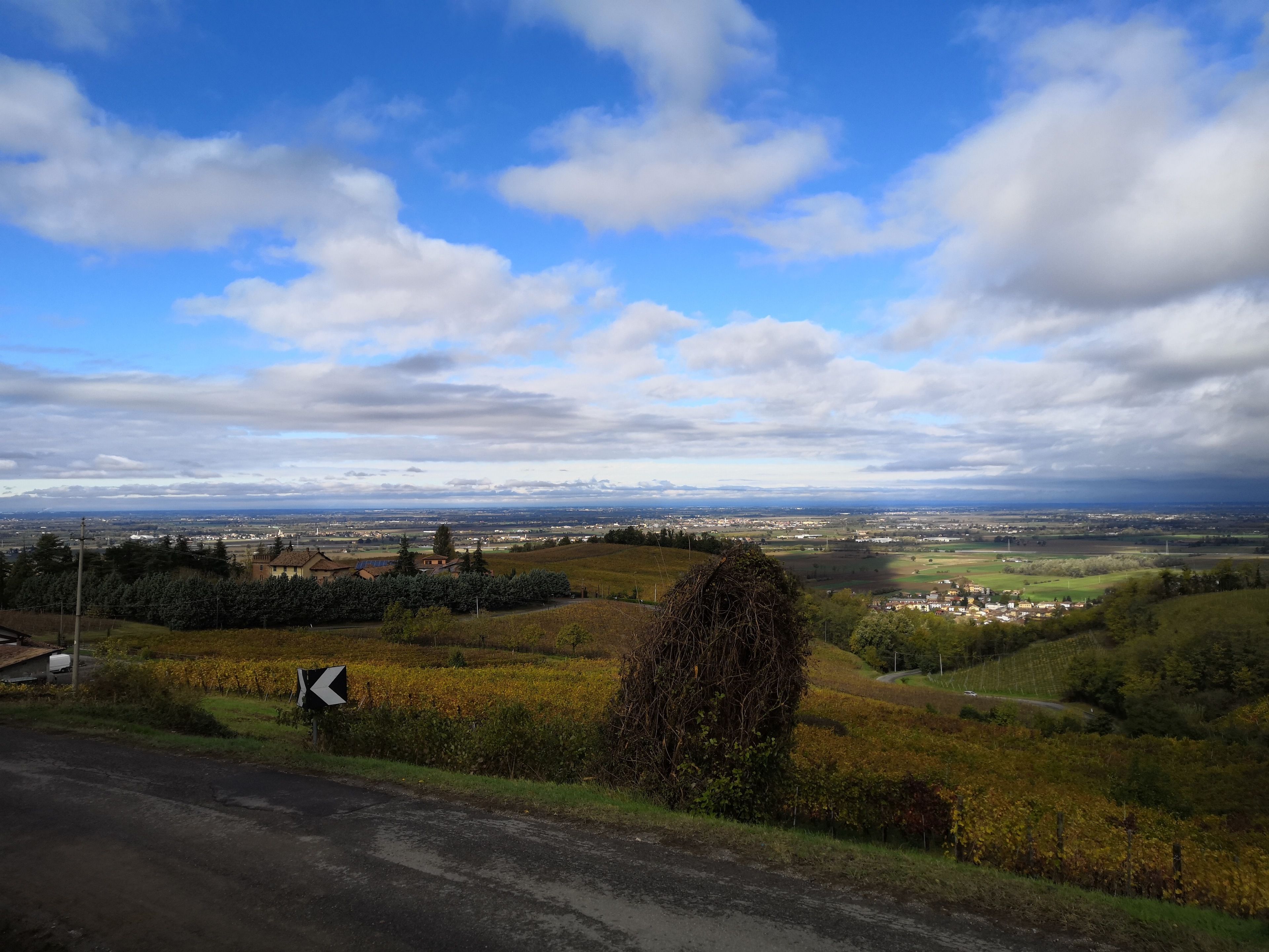 A rural landscape with a road, fields, and distant buildings under a partly cloudy sky.