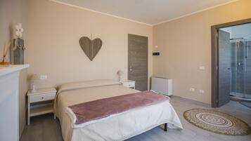 A bedroom with a bed, bedside tables, a nightstand, a decorative heart on the wall, and a shower area visible through an open door.