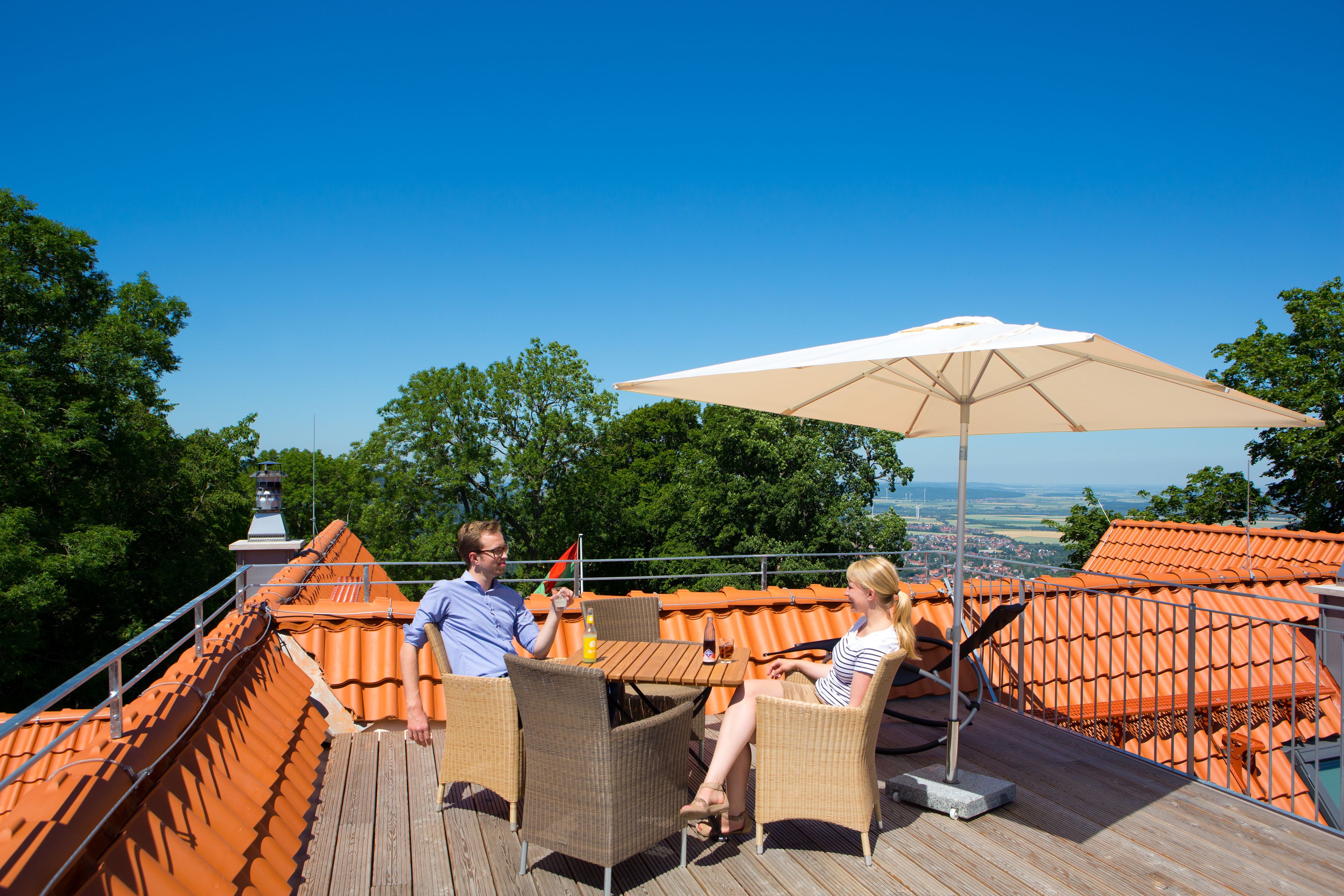 Una pareja sentada en una mesa bajo un gran paraguas en una terraza de madera con techo naranja, con vistas a un paisaje pintoresco.