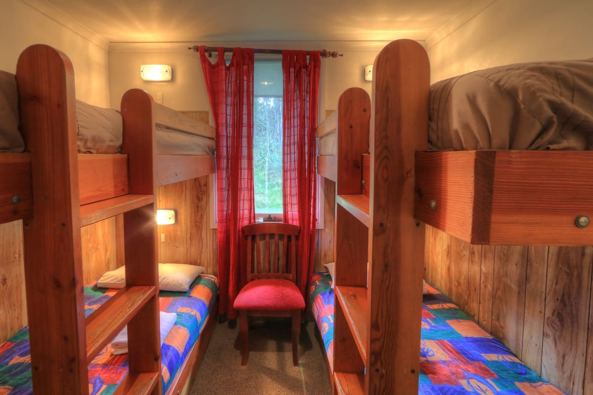 Una habitación pequeña con literas, una silla de madera en el centro y una ventana con cortinas rojas.