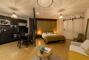 Quarto de hotel compacto com cozinha americana, cama com colcha verde, área de refeições com mesa e cadeiras, e sala de estar com sofá.