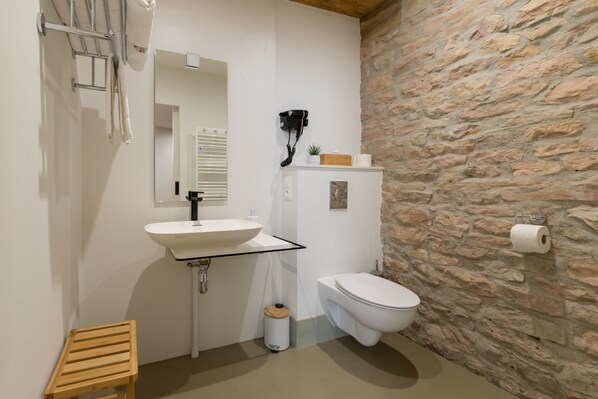A bathroom with a stone wall, a white sink, a toilet, a wooden stool, and a hairdryer mounted on the wall. - Appart Hotel Eugenie (Louhans)