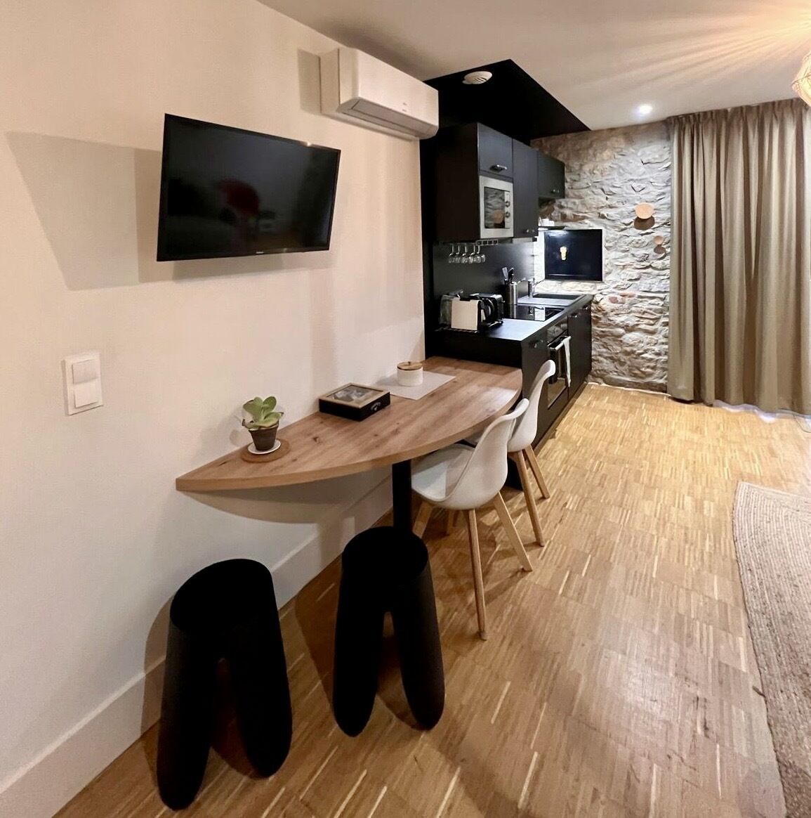 Una cocina moderna con una pared de acento de piedra, un televisor de pantalla plana montado sobre la encimera y una zona de comedor con mesa de madera y sillas blancas.
