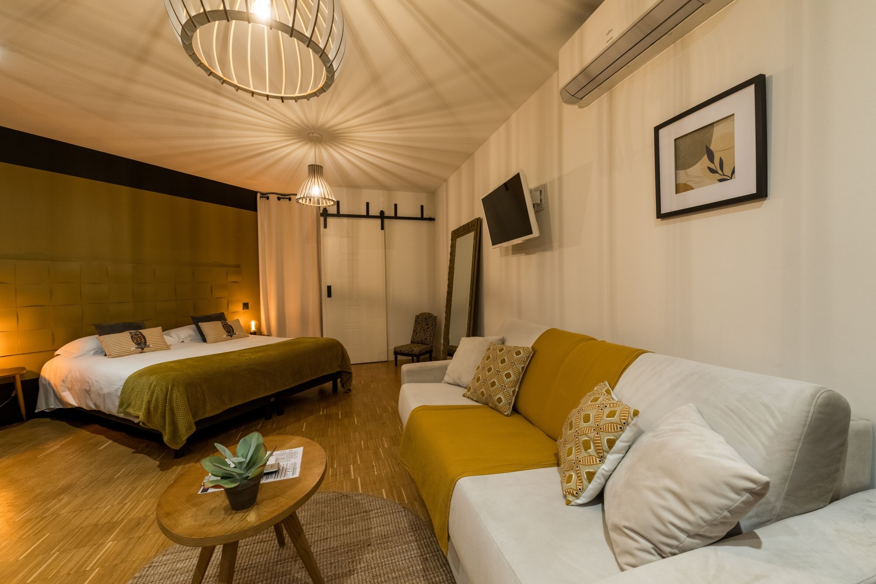 Ein Hotelzimmer mit Bett, Sofa, einem kleinen Tisch mit Pflanze, einem an der Wand montierten Fernseher und einer Deckenleuchte.