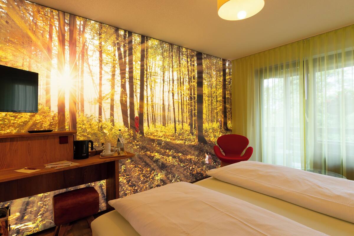 A hotel room with a bed, a desk with a coffee maker, a red chair, and a large forest mural on the wall.