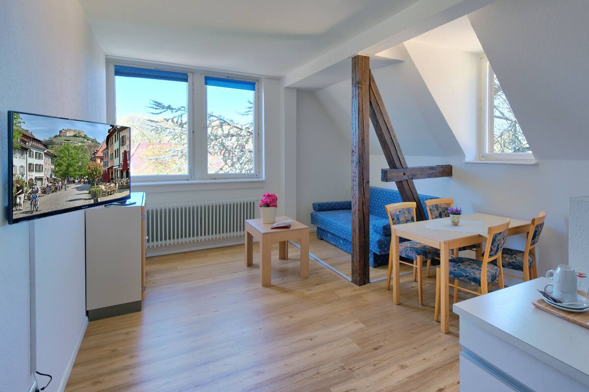 A modern apartment with a loft-style room, large windows, a dining area, and a TV mounted on the wall.