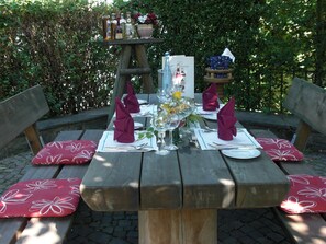 Et udendørs picnicbord dækket med røde blomstrede puder, pænt arrangerede tallerkener, bestik og vinglas.