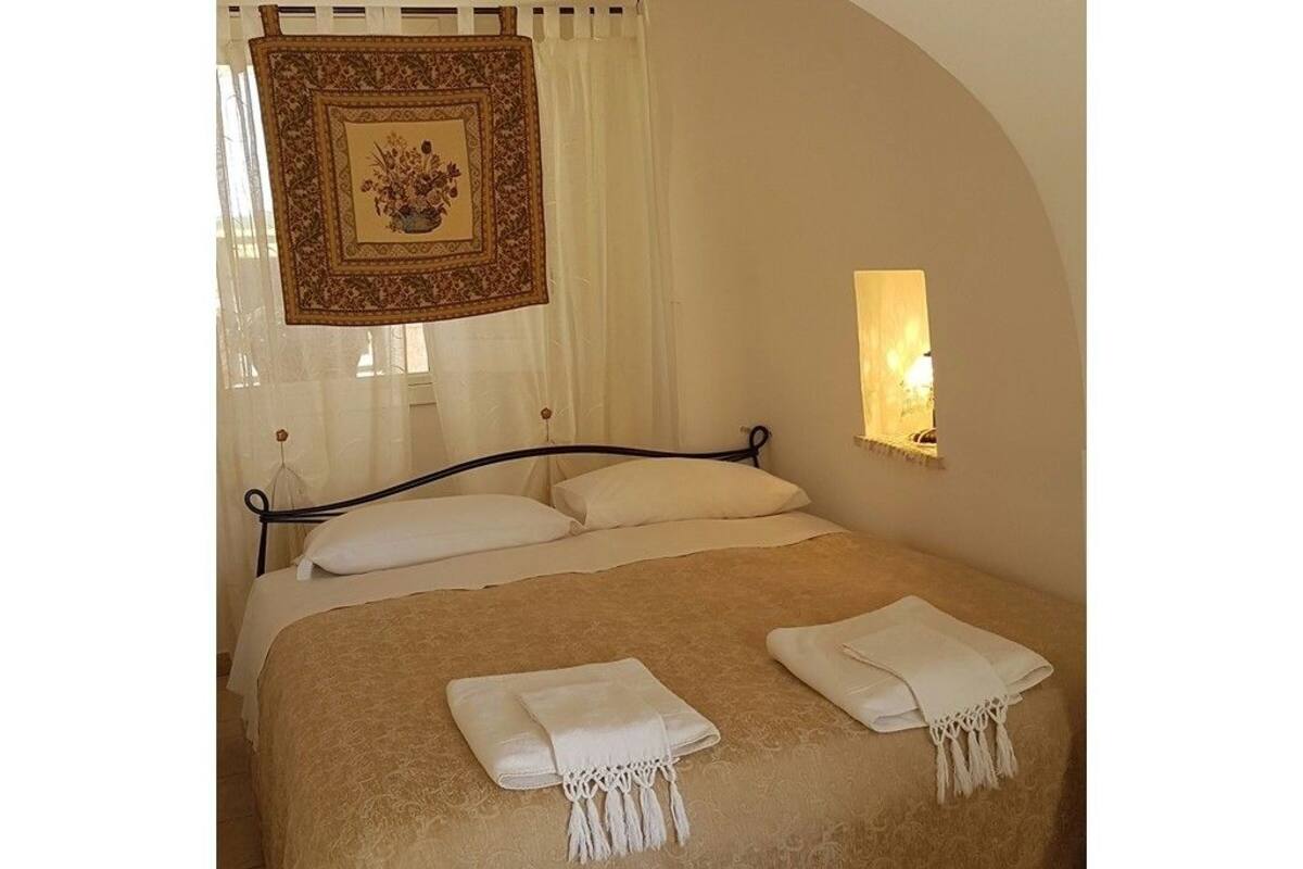 Uma cama bem arrumada com lençóis brancos e duas toalhas dobradas, um objeto decorativo pendurado acima da cama, e uma luminária fixada na parede.