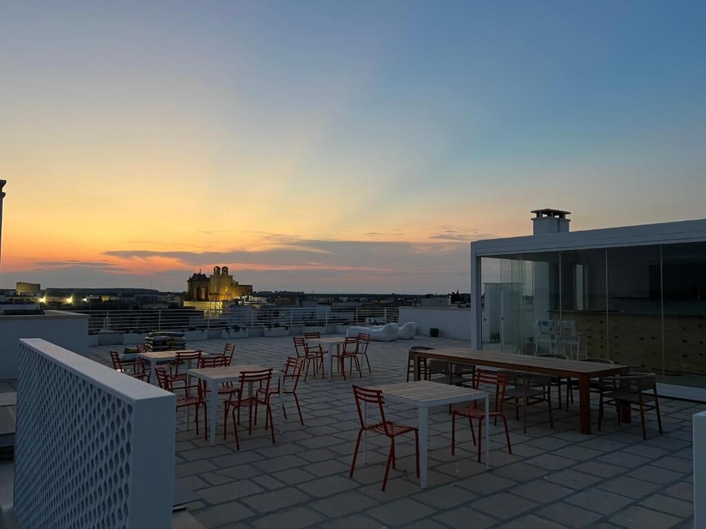 A rooftop terrace with outdoor seating, a glass wall, and a view of a cityscape at sunset.