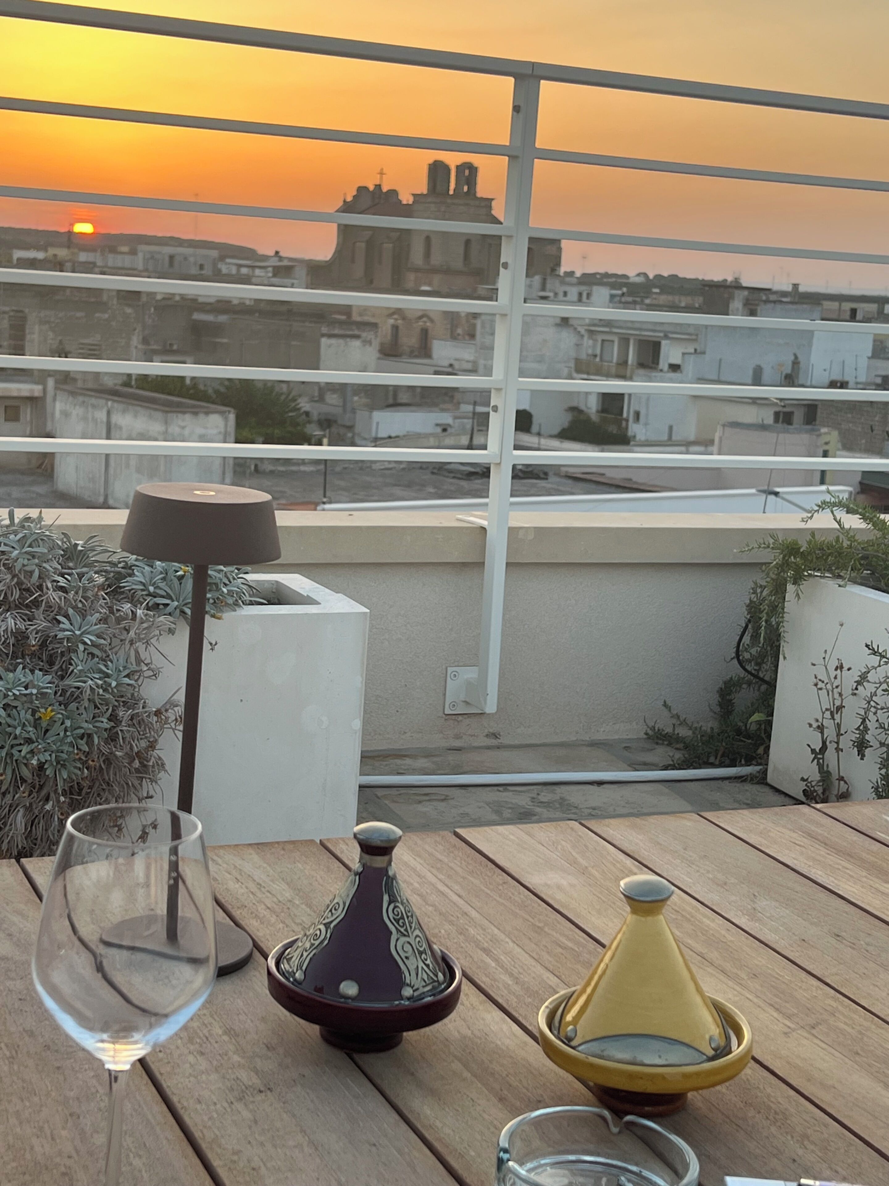 Et bord med et vinglas, en dekorativ flaske og en tekedel på en balkon med udsigt over en by ved solnedgang.