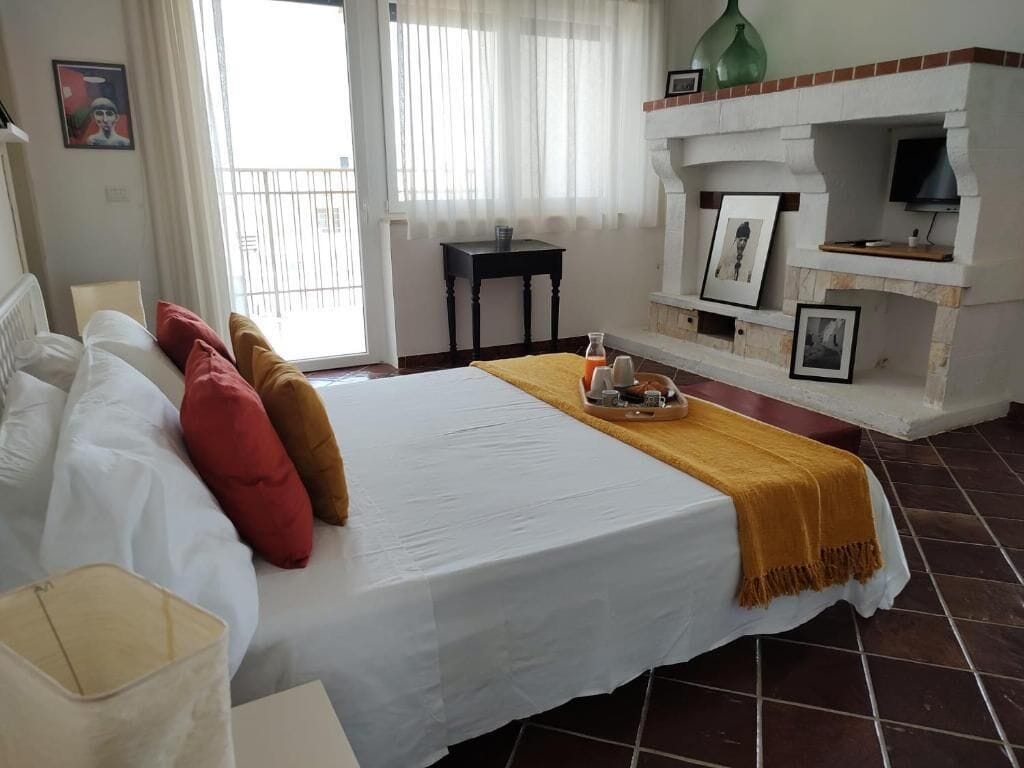 A hotel room with a bed, a small table, a chair, a fireplace, and a balcony.