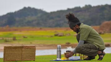 A person preparing food outdoors with a basket, a thermos, and a jar on a mat.