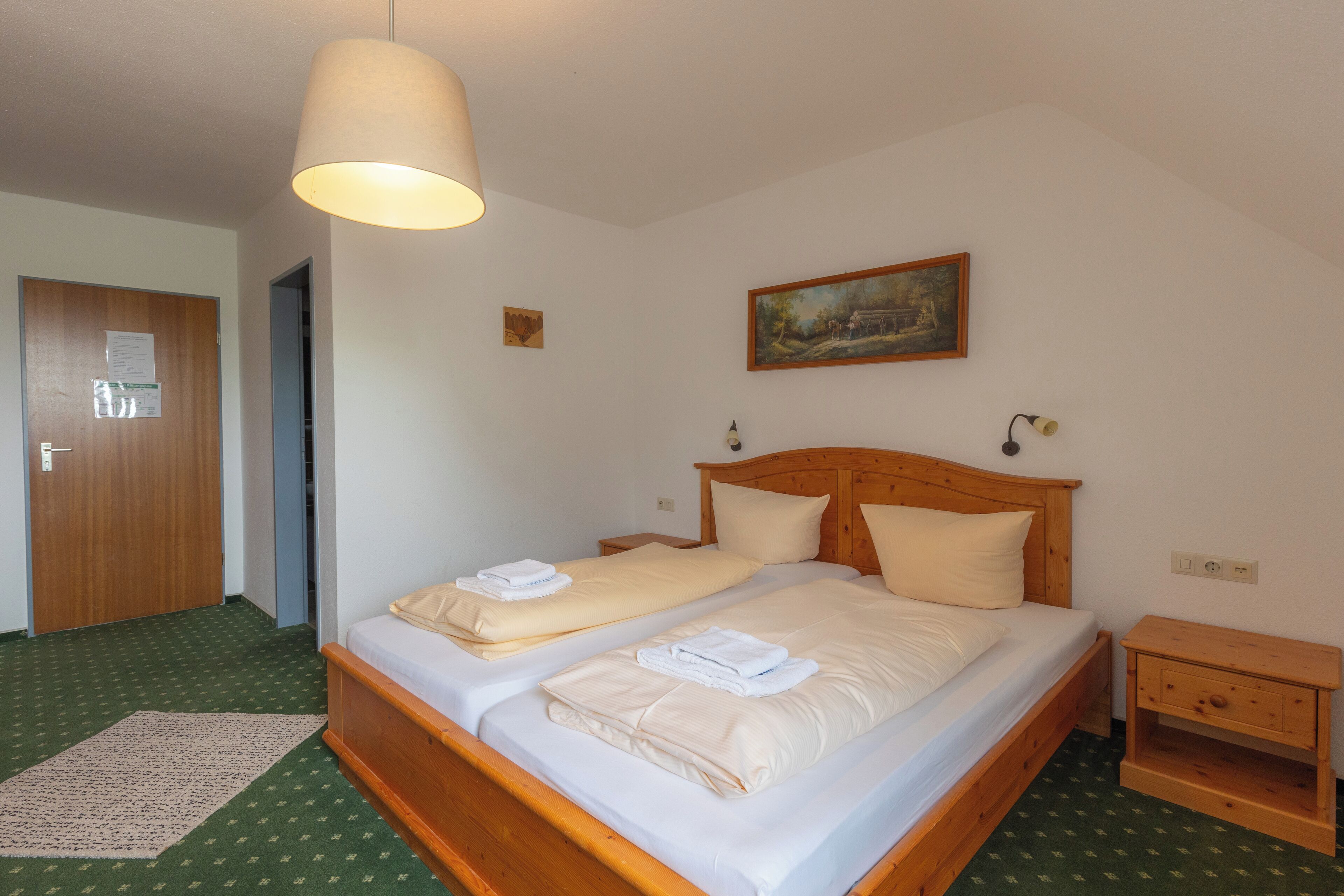 A hotel room with a double bed, wooden headboard, two bedside tables, a small wooden nightstand, a hanging lamp, a painting on the wall, and a green patterned carpet.