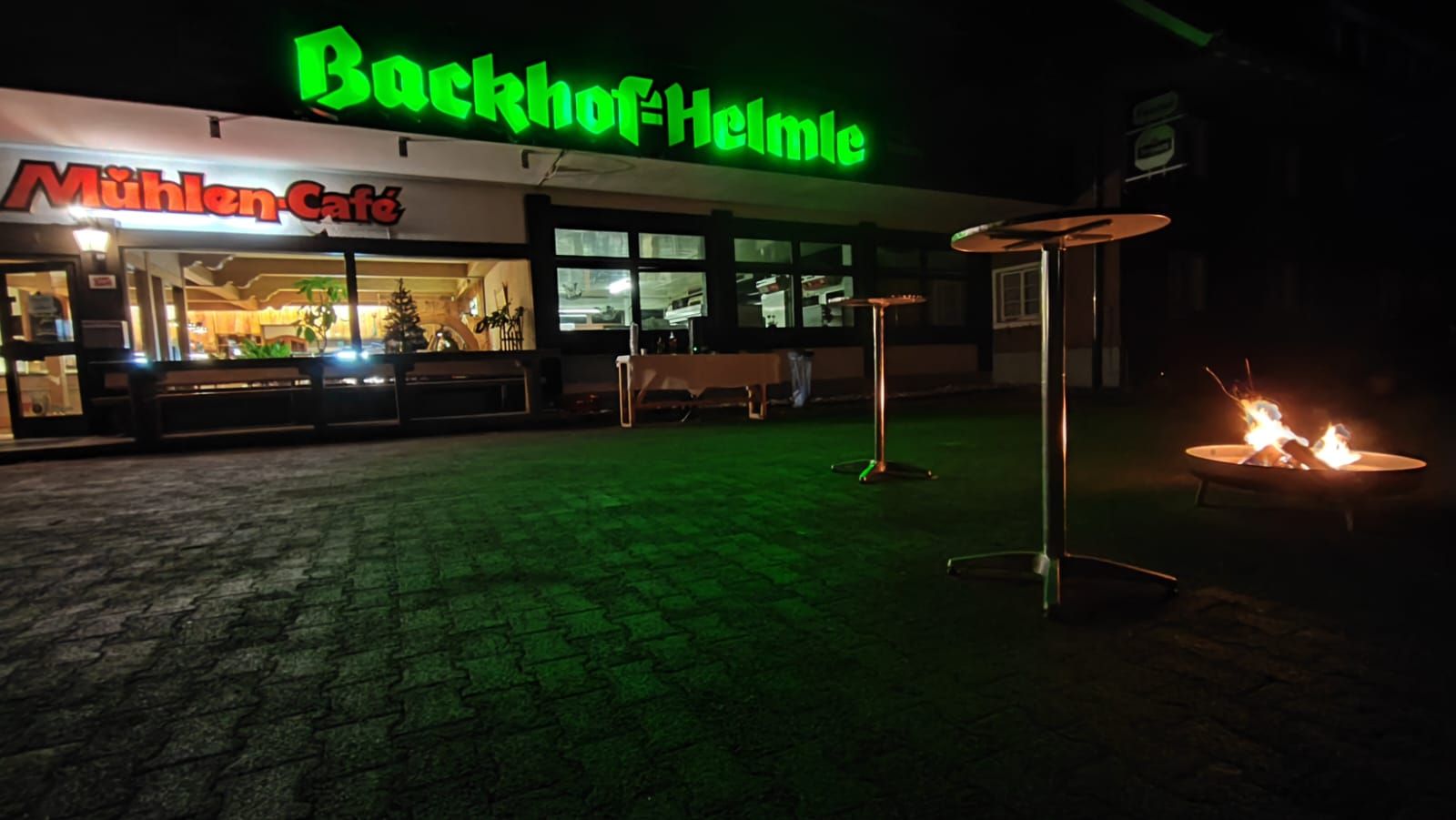 Een gebouw met een neonbord waarop 'Backhof Heimle' en 'Mühlen Cafe' staat vermeld. Er zijn buitentafels en een vuurkorf.
