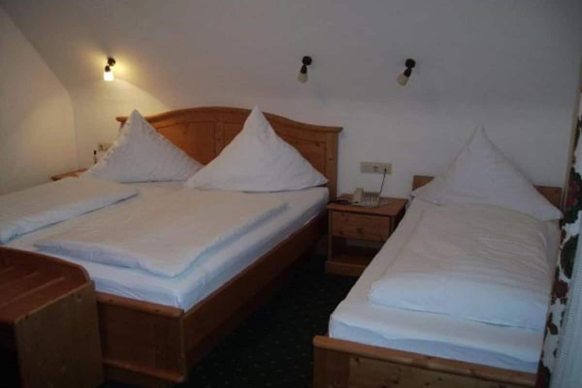 Una habitación con dos camas de madera, cada una con cabecero de madera y ropa de cama blanca. Hay mesitas de noche y una lámpara fijada a la pared.