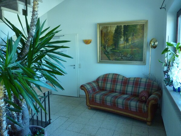 A living room with a plaid sofa, a painting of a forest, a potted plant, and a door. - Gasthaus Backhof-Helmle  (Breitnau)