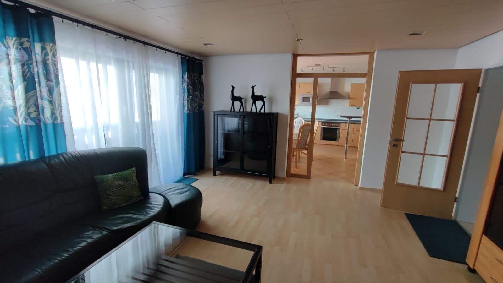 Una sala de estar con un sofá negro, una mesa de centro de vidrio y un mueble para televisión. Se ve una cocina a través de un vano abierto.