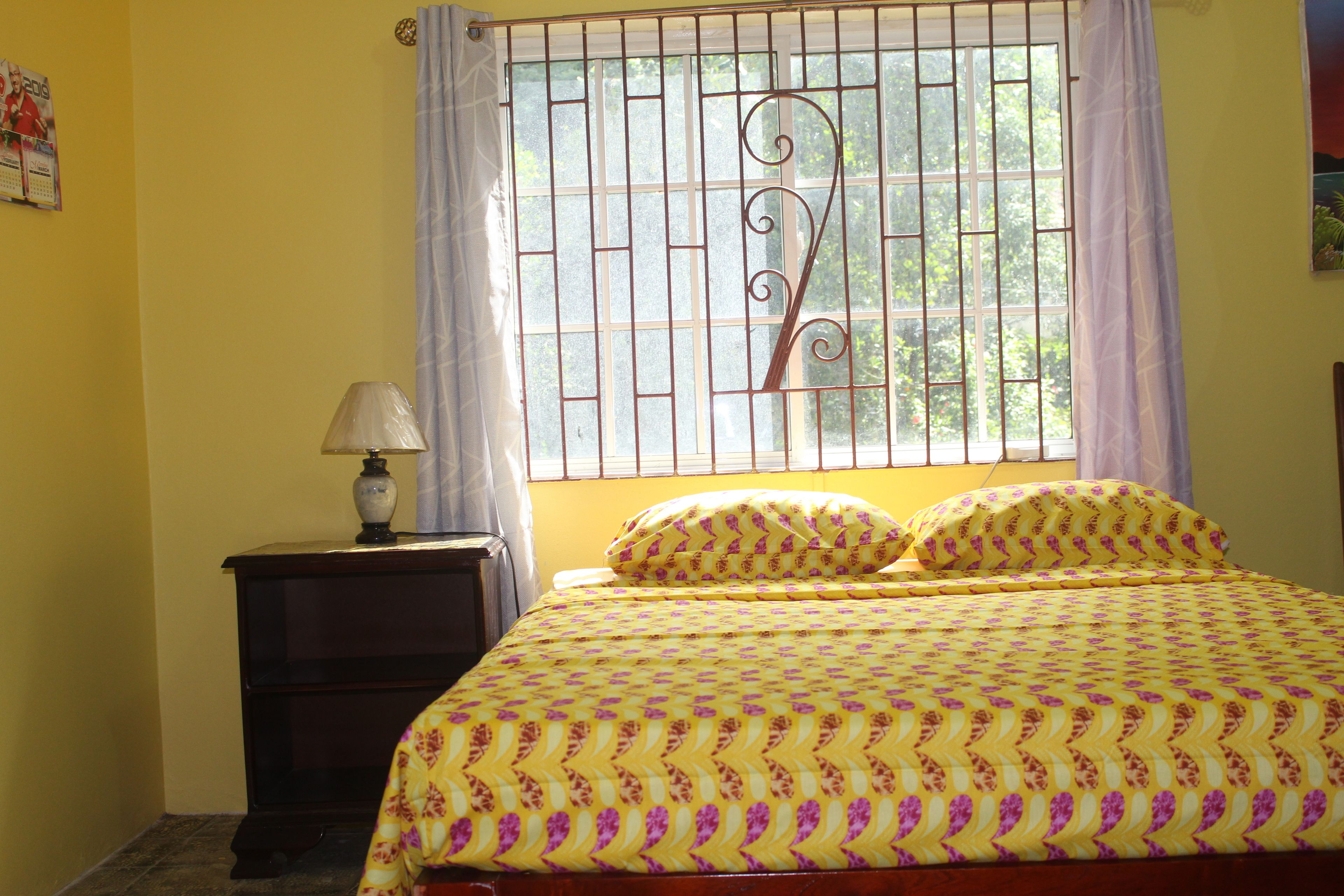 A bedroom with a bed, a nightstand, a lamp, and a window with bars.