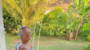 An elderly person seated on a chair in a covered outdoor area with a stone floor, surrounded by potted plants and greenery. - ritashomeawayfromhome (Ocho Rios)