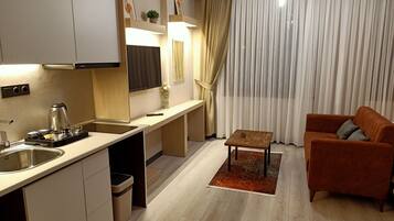 A modern kitchen with a built-in oven, a coffee maker, and a sink. There is a small table with a vase of flowers and a remote control. The living area has a brown sofa and a rug.