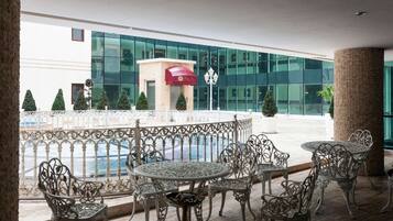 A hotel lobby with ornate metal chairs and tables, a red awning, and a view of a modern building with glass windows.