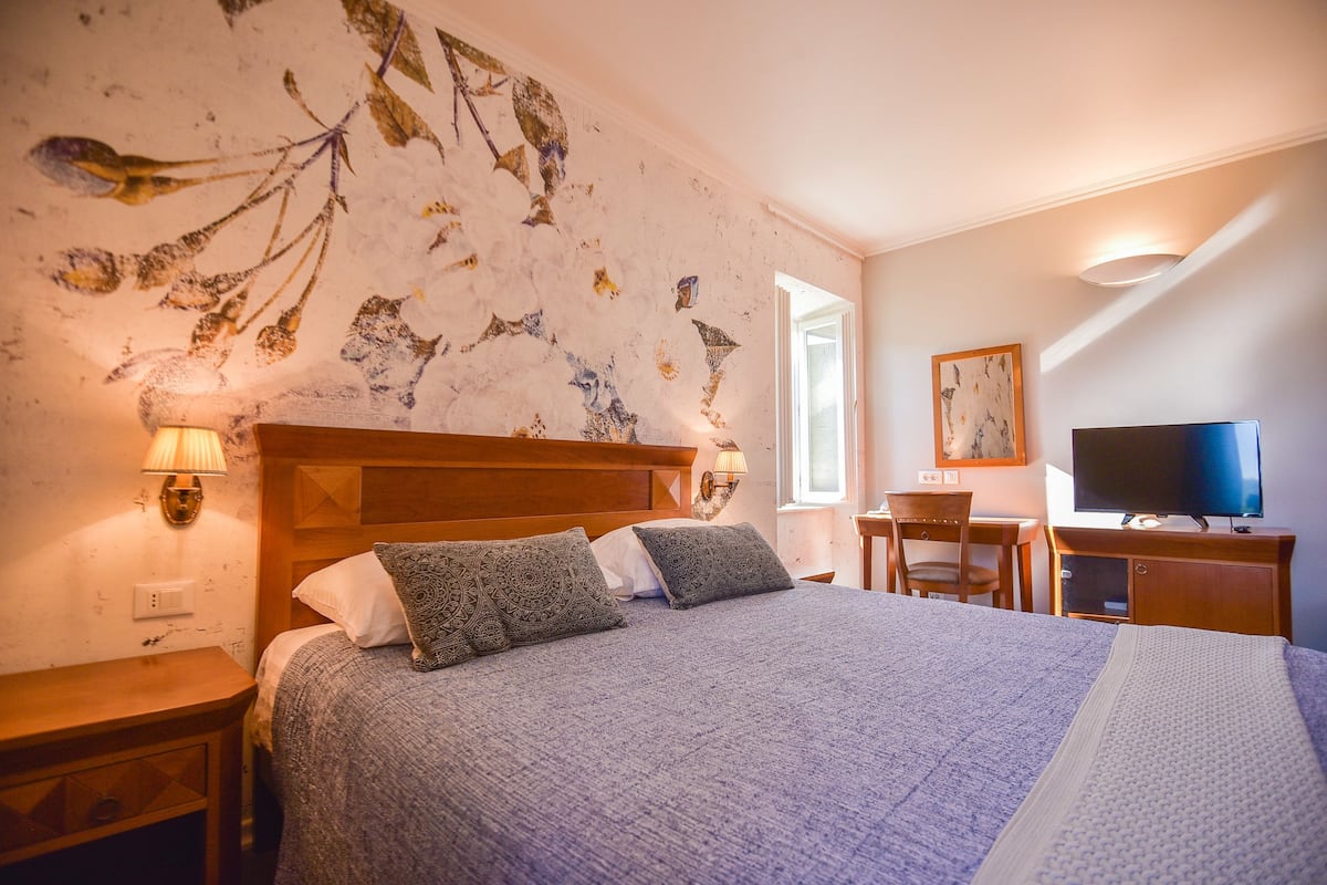 A bedroom with a large bed, a desk, a chair, a television, and a wall with a floral mural.