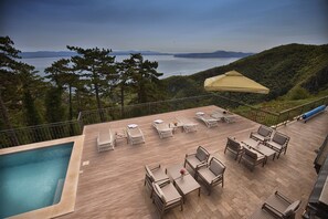 A rooftop pool area with lounge chairs and a table, overlooking a body of water and hills. - Hotel Draga di Lovrana (Lovran)