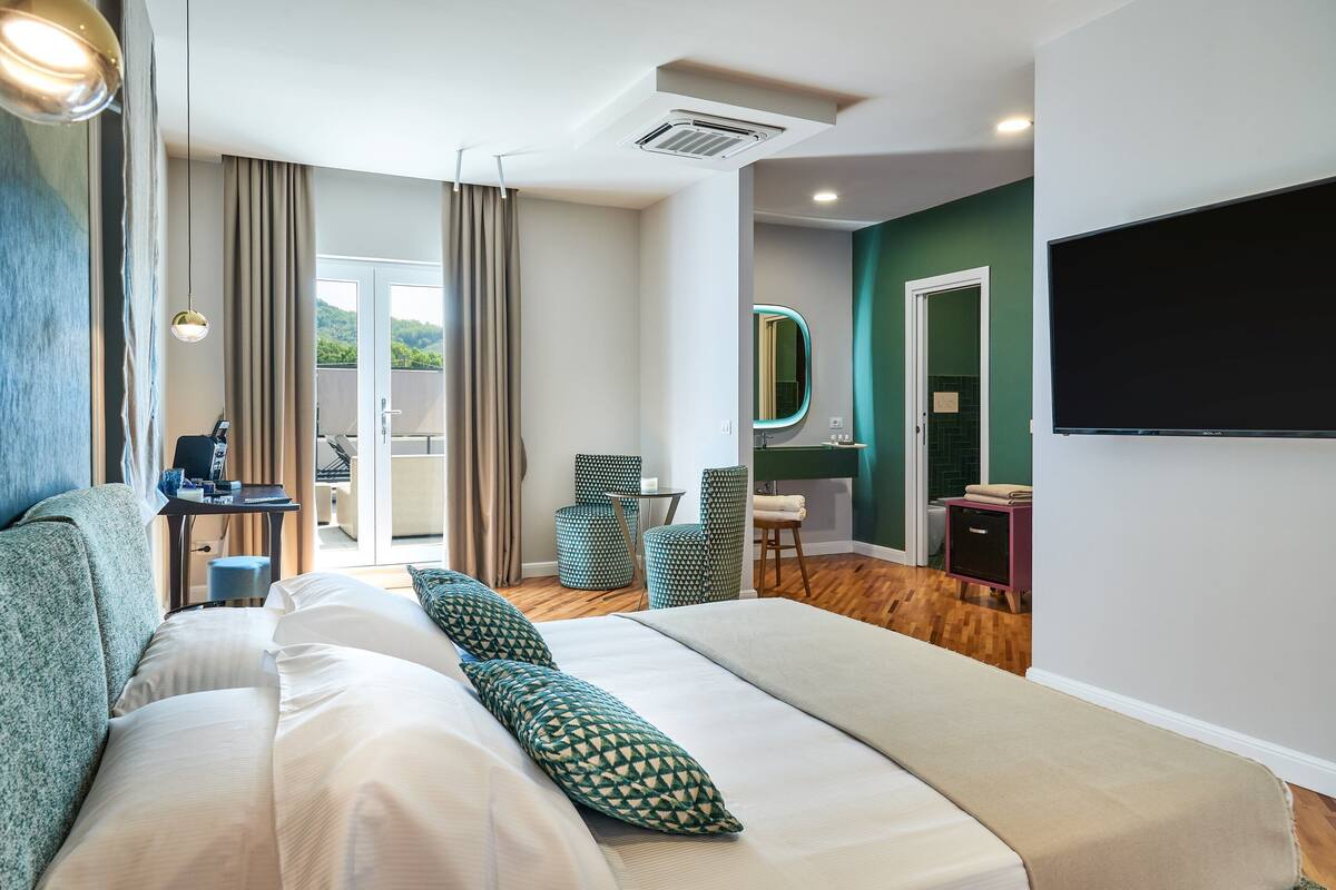 A modern hotel room with a large bed, a desk with a computer, a chair, a small table, a mirror, and a TV.