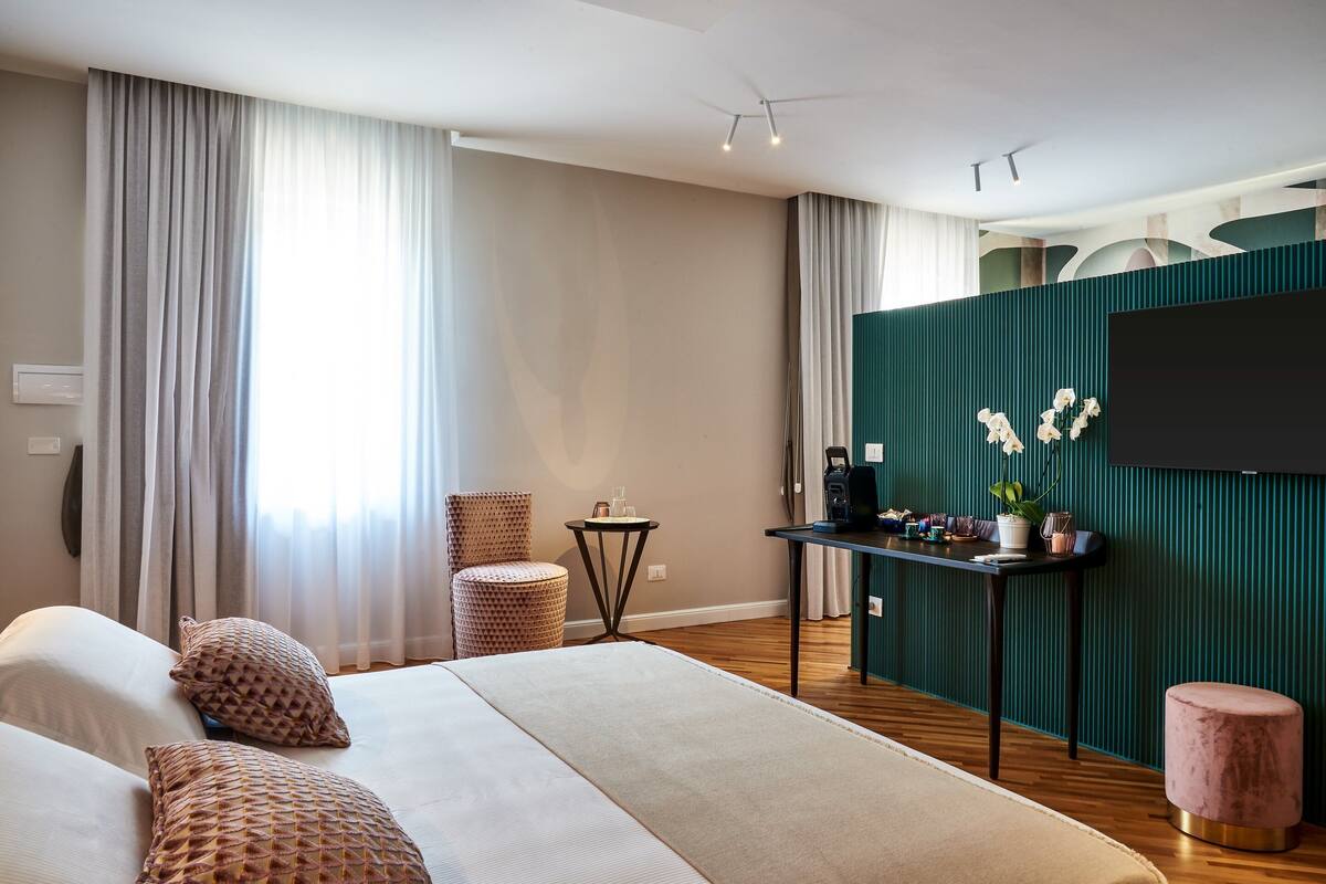 A modern hotel room with a large bed, a sitting area with a chair and a small table, a flat-screen TV mounted on a green wall, and a large window with curtains.