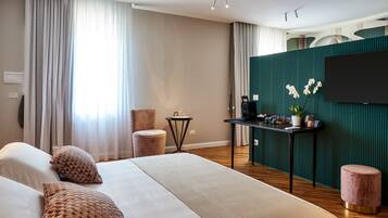 A modern hotel room with a large bed, a sitting area with a chair and a small table, a flat-screen TV mounted on a green wall, and a large window with curtains.