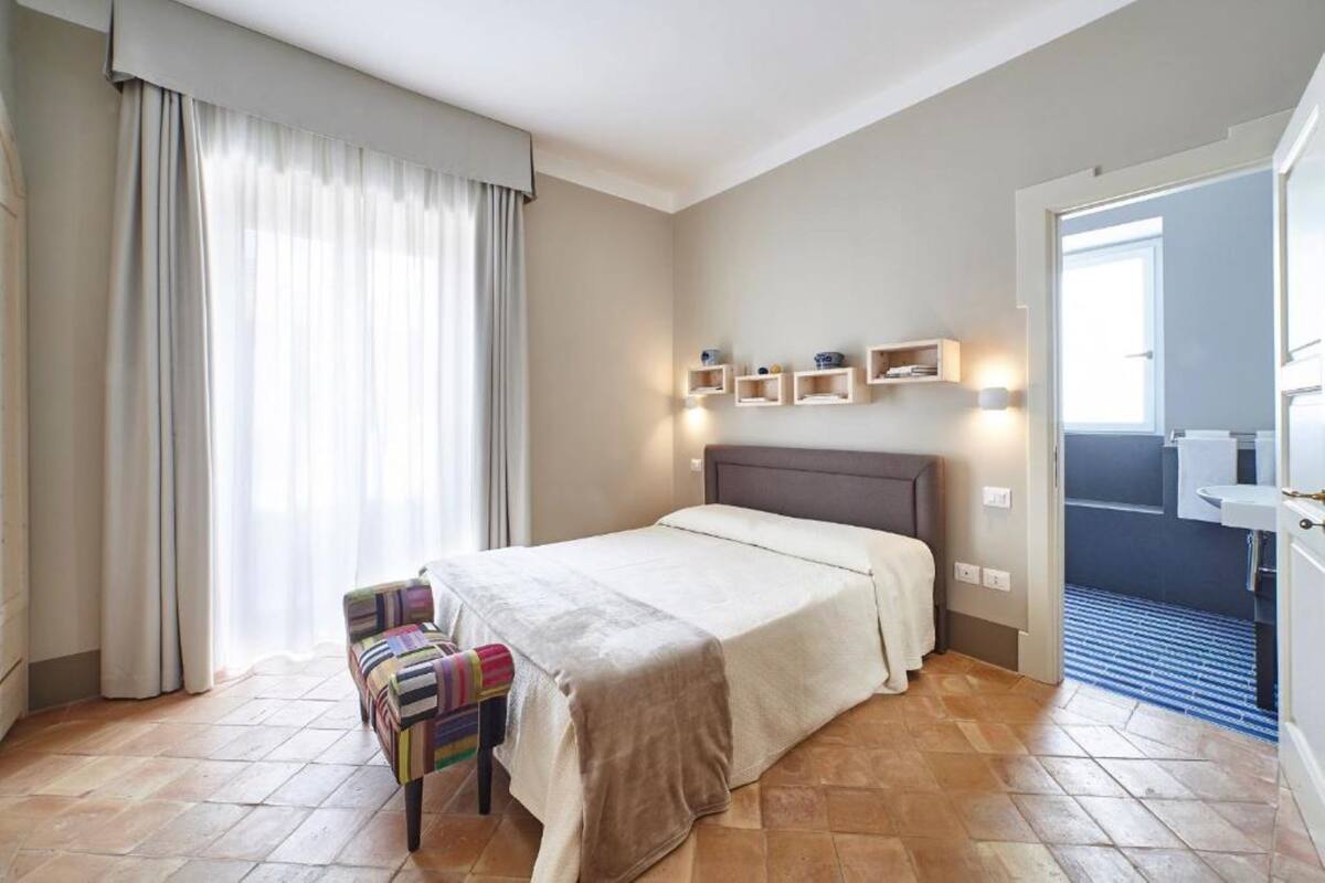 A hotel room with a bed, a chair, a bedside table, and a bathroom visible through an open door.