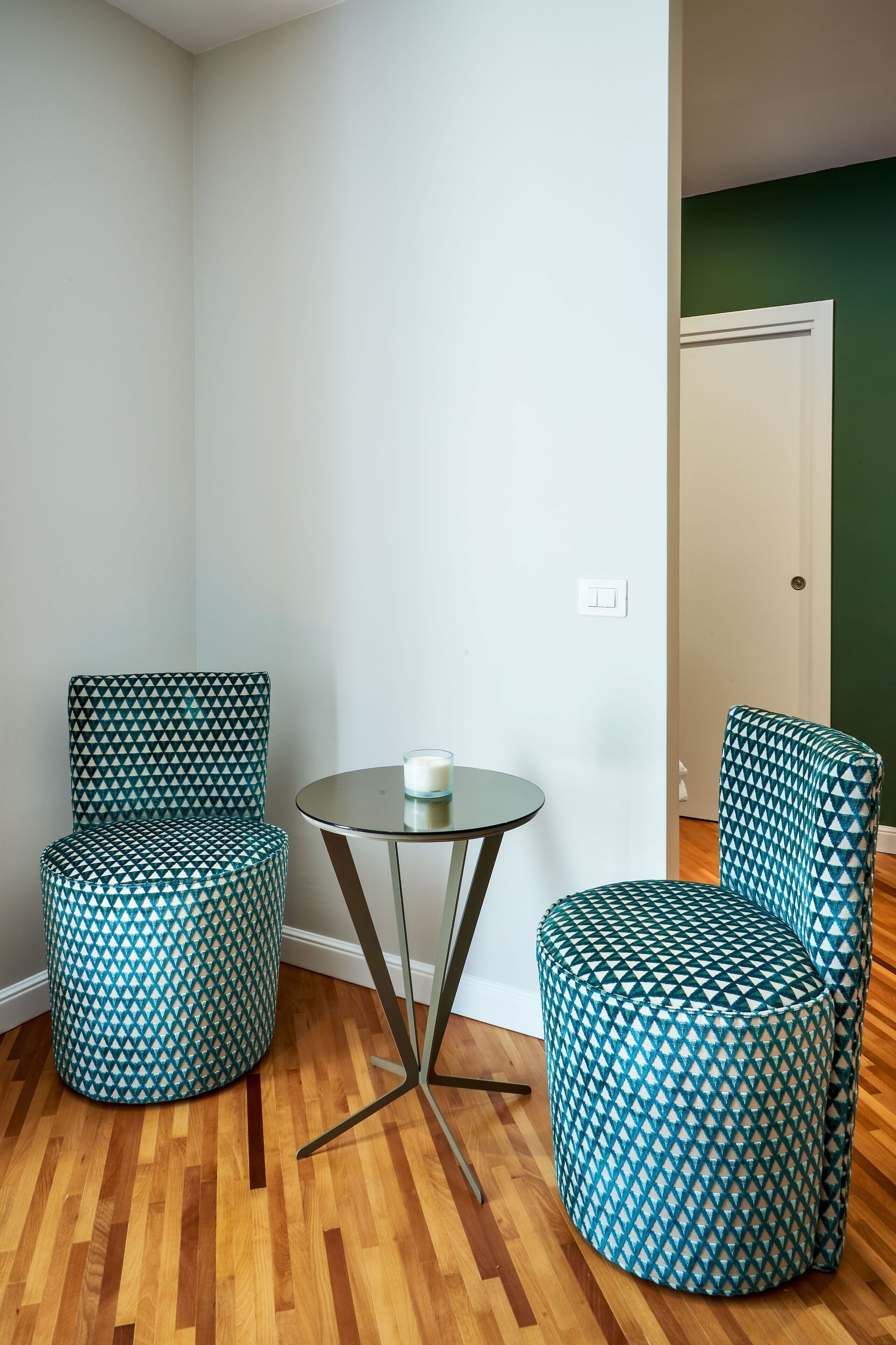 Habitación de esquina con dos sillones estampados, una mesita redonda y una puerta blanca.