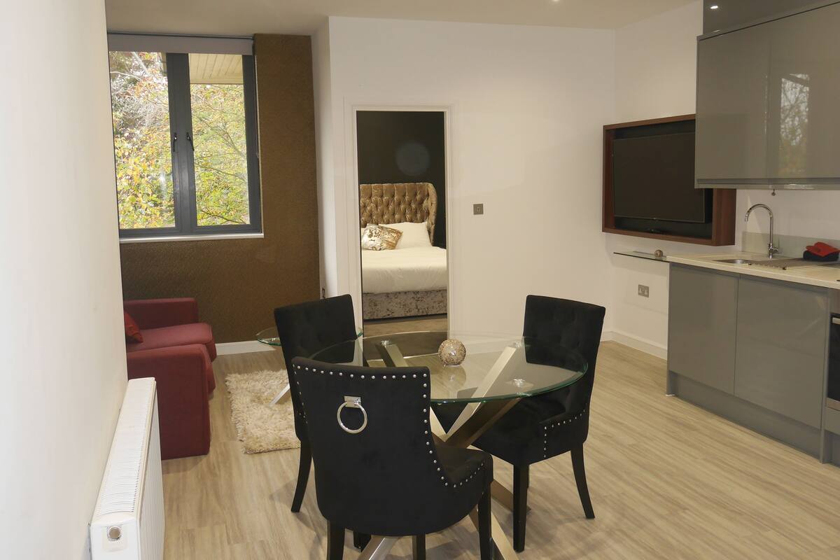 A compact kitchen with a dining area, a TV mounted above the sink, and a view of a bedroom through an open door.