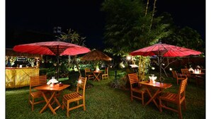 Un espacio exterior para comer con mesas, sillas y una zona de bar, todo bajo sombrillas rojas.