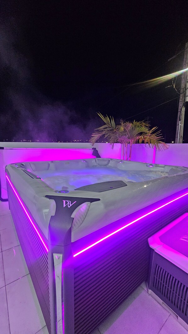 Uma banheira de hidromassagem com iluminação LED roxa, rodeada por plantas de palmeira, em um terraço no topo do prédio.