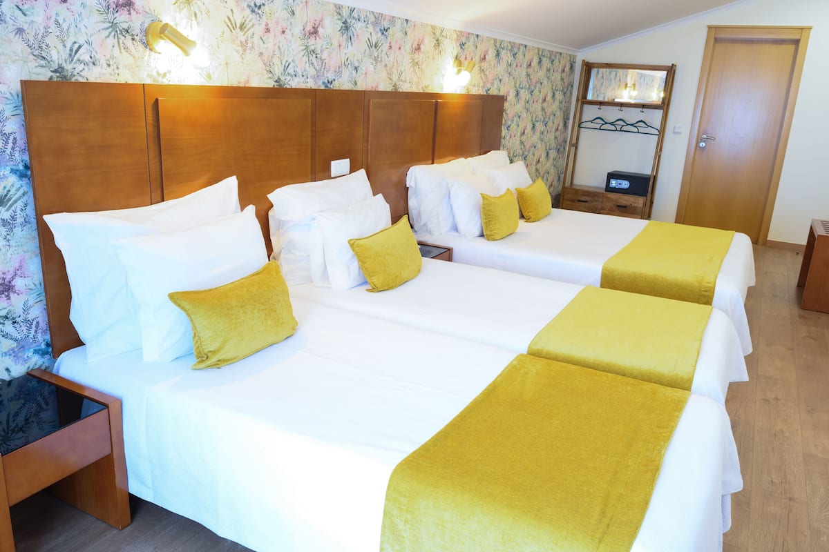 A hotel room with two beds, each with white bedding and yellow pillows. There is a wooden headboard, a nightstand, and a small wooden cabinet. The room has floral wallpaper and a wooden door.