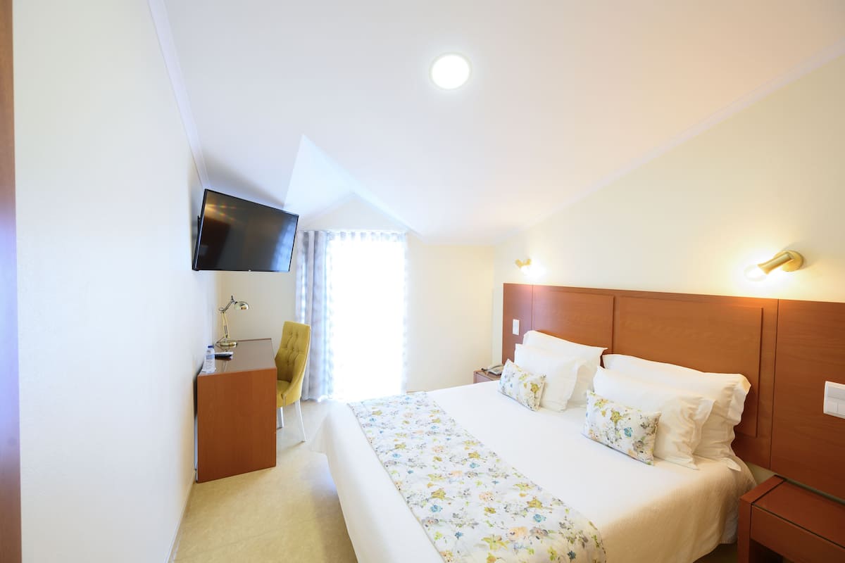 A hotel room with a bed, bedside tables, a desk with a chair, a flat-screen TV mounted on the wall, and a window with curtains.