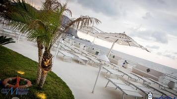 Zona de relax a la platja amb gandules blanques i ombrel·les, una palmera i un bou salvavides sobre la gespa.
